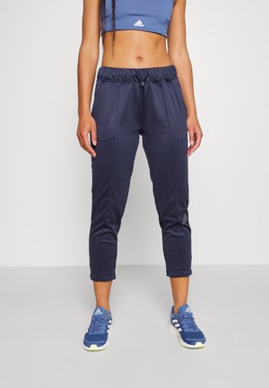 Nainen pukeutuneena tummansinisiin lyhyisiin verkkarit, siniseen Adidas-urheiluliiviin ja sinisiin Adidas-tennareihin seisoo yksinkertaista vaaleaa taustaa vasten.