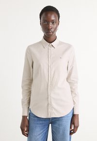 Light beige long-sleeve shirt with a collar, front buttons, and a small embroidered logo. Paired with blue jeans, shown in a neutral background.