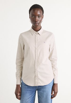 Lys beige langermet skjorte med krage, frontknapper og en liten brodert logo. Kombinert med blå jeans, vist på en nøytral bakgrunn.