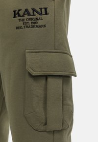 Olijfgroene cargobroek van zachte stof; beschikt over een klepzak aan de zijkant met stiksels en geborduurd logo-detail.