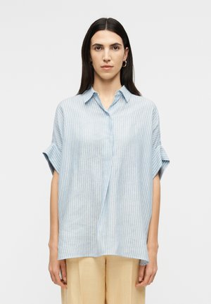 Kobieta o długich ciemnych włosach, ubrana w jasnoniebieską koszulę w pionowe paski z krótkim rękawem oraz beżowe spodnie, stojąca na tle jednolitego jasnego tła.