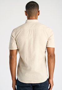 Man met kort haar in een beige kortje met mouwen en donkere broek, staande met een witte achtergrond en ontspannen armen aan zijn zij.