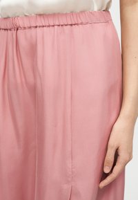 Κοντινό πλάνο ενός ατόμου που φοράει μεταξένιες ροζ παντελόνες με ελαστική μέση, δείχνοντας τη ραφή στο πλάι και το δεξί του χέρι που ακουμπά δίπλα στο παντελόνι.