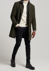 Manteau en laine mélangée olive sur une doudoune noire, col roulé crème et jean noir, associé à des bottes noires à lacets.