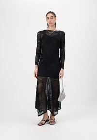 Svart spetsklänning med långa ärmar, asymmetrisk nederkant, genomskinlig textur och geometriska mönster, i kombination med en liten metallisk handväska.