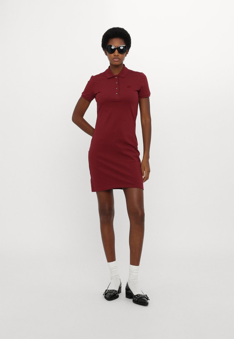 Femme portant une robe polo bordeaux à manches courtes, des lunettes de soleil noires, des chaussettes blanches et des chaussures à talons noires, se tenant devant un fond blanc uni.