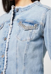 Chaqueta de mezclilla azul claro con bordes deshilachados y brillantitos decorativos en la tapa del bolsillo superior, que lleva puesta una persona.