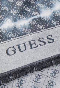 Blå og grå skjerf med et diamantmønster som har et gjentatt logo. Ferdige kanter med frynser. "GUESS" vises tydelig i svart.