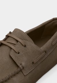 Μοκασίνι από σουέντ δέρμα σε απόχρωση ταούπ, με ραμμένη μύτη, λεπτομέρεια κορδονιών και μαλακό επενδυμένο εσωτερικό.