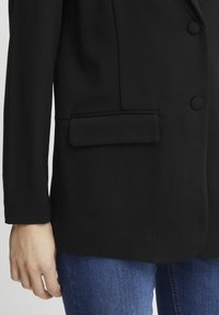 Svart kavaj med enkelknäppning, slät yta, två framfickor och spetsig revers. Bärs över blå jeans.