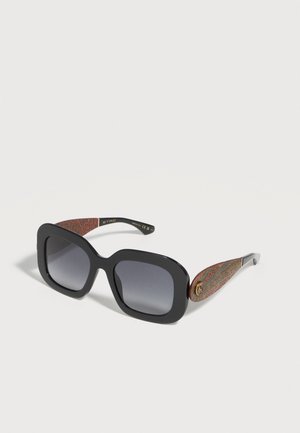 Lunettes de soleil carrées oversize noires avec verres dégradés foncés et branches multicolores texturées avec un petit logo rond doré.