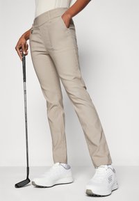 Beige golfnadrág sima textúrával, oldalzsebekkel és lapos elülső kialakítással, fehér sportcipőkkel párosítva.