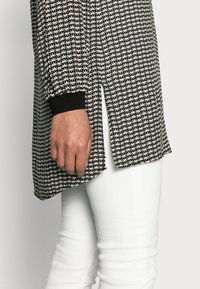Túnica em padrão preto e branco com acentos laranja, apresentando uma ligeira fenda lateral e punhos pretos, combinada com calças brancas ajustadas.
