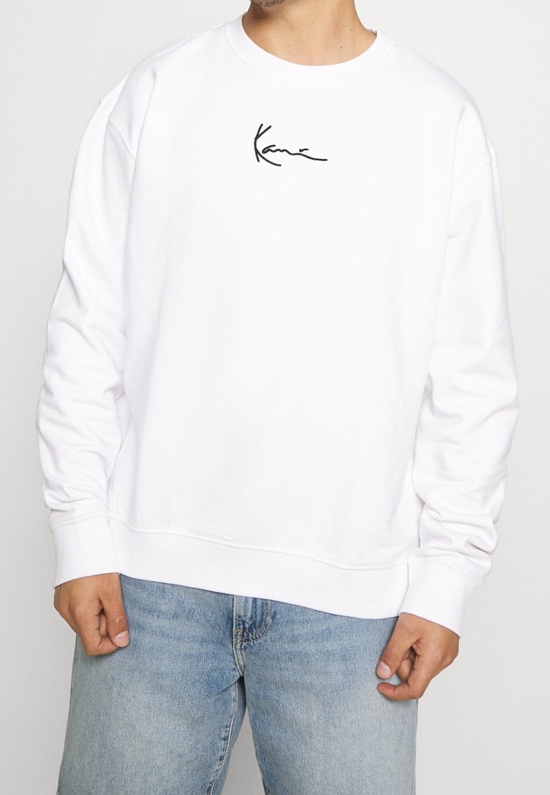 Sudadera blanca de algodón con puños y dobladillo de canalé, con un logo distintivo negro en el pecho. Combinada con vaqueros de mezclilla azul claro.