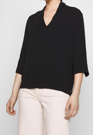 Femme portant un chemisier noir à col en V avec des manches trois-quarts et un pantalon beige clair, debout devant un fond uni.