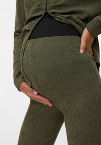 Zwangere persoon in een textuurstof olijfgroen zwangerschapsbroek en bovenstuk, die zachtjes hun babybuik met beide handen vasthoudt.