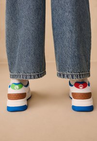 Baskets blanches avec des accents bleus, ornées de logos "L" verts et "M" rouges sur les talons et des détails en motif brique. Portées avec un pantalon en denim bleu.