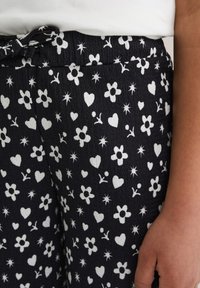 Black fabric pants with white floral and heart patterns, featuring a textured finish and an elastic waistband with adjustable tie detail.