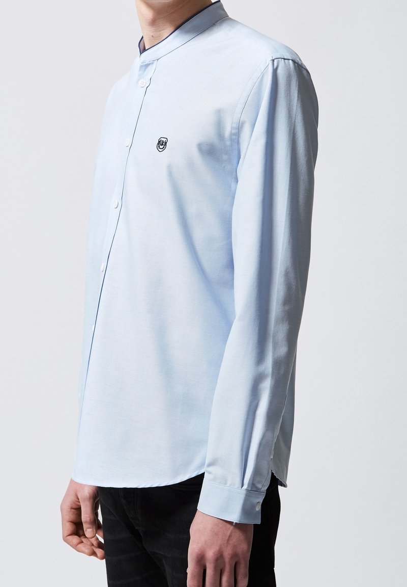 Chemise bleu clair à manches longues avec col mandarin et fermeture à boutons, comportant un petit logo brodé sur la poitrine.