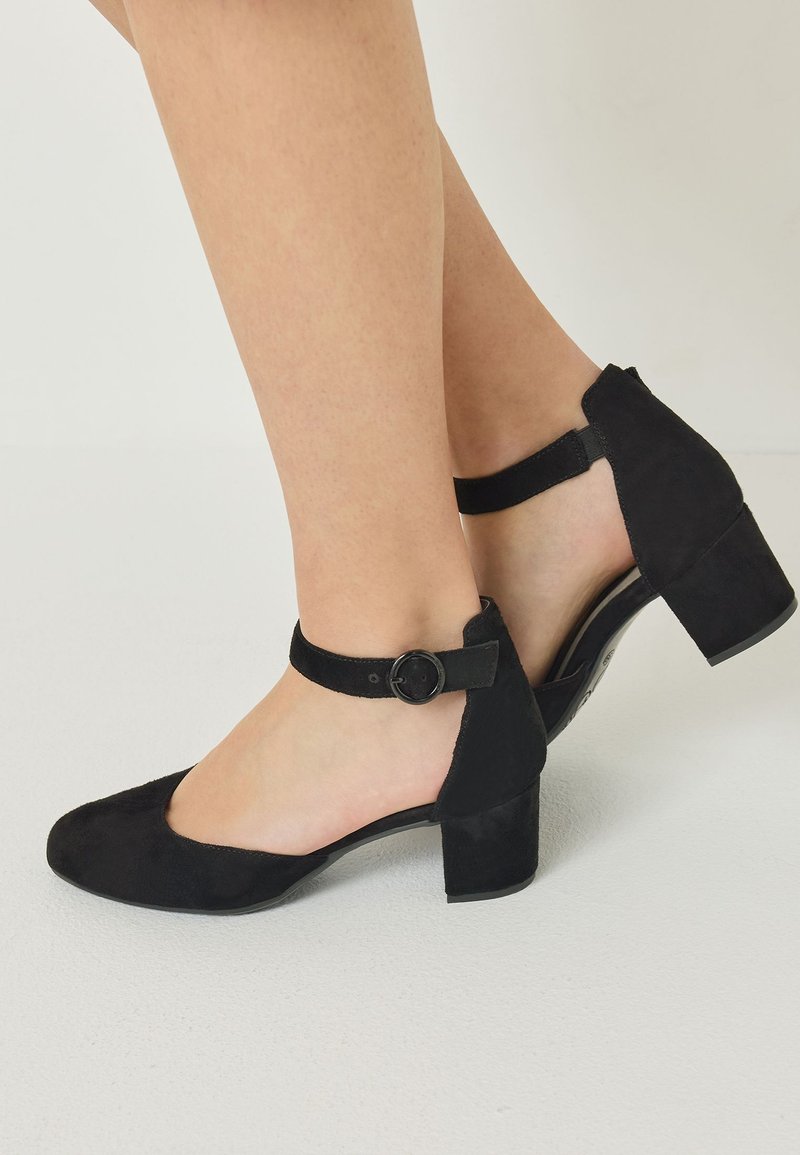 Chaussures Mary Jane en daim noir avec brides à boucle ronde et talons blocs portées pieds nus sur un fond clair uni.