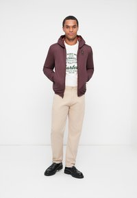 Chaqueta ligera con capucha color marrón, pantalones beiges, camiseta gráfica blanca y zapatos negros. Textura suave con cremallera y acentos de logo.