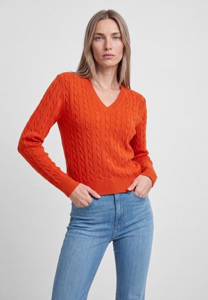 Frau mit langen blonden Haaren, die einen orangefarbenen gestrickten V-Ausschnittpullover und hellblaue Jeans trägt, steht mit den Händen in den Taschen.