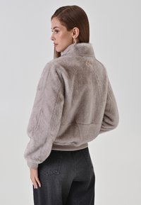 Giacca in pelliccia sintetica di morbido color taupe con colletto alto, polsini a costine e dettaglio del logo sul retro. Abbinata a jeans neri a vita alta.
