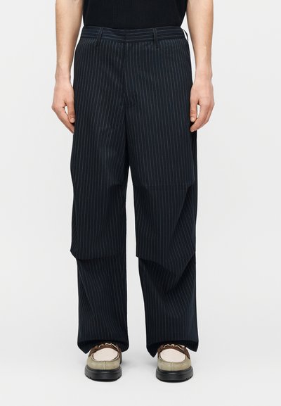 Παντελόνι σε στυλ wide-leg με ναυτική πλαινή ρίγα, φορεμένο με μπεζ loafers που διαθέτουν χρυσή αλυσίδα και μαύρο τοπ ορατό στη μέση.