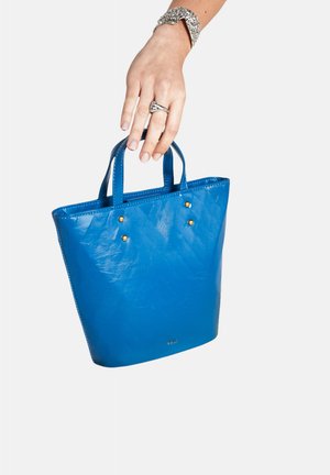 Blauwe leren draagtas met kleine gouden studs, korte handvatten en een glanzende textuur, wordt vastgehouden door een hand.