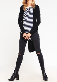 Svart långärmad cardigan över en vit och marinrandig skjorta, kombinerad med svarta slitna skinny jeans och ankle boots. Enkla smycken är synliga.