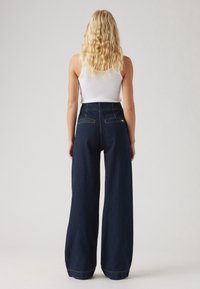 Mujer con cabello rubio largo y ondulado que lleva una blusa sin mangas blanca y unos pantalones de mezclilla oscuros de cintura alta y pierna ancha, de pie contra un fondo sencillo.