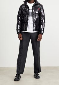 Osoba w błyszczącej czarnej kurtce puchowej, białym t-shircie z grafiką, czarnych dopasowanych spodniach i błyszczących czarnych loafersach stoi na jasnej podłodze.