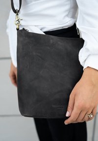 Schwarze Leder-Crossbody-Tasche mit glatter Textur, ausgestattet mit einem Reißverschluss, verstellbarem Tragegurt und geprägtem Markenlogo.