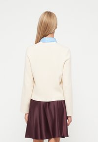 Kremfarget strikkegenser med lange ermer og en blå skjorte synlig under, brukt med et brunt satinflared skjørt.