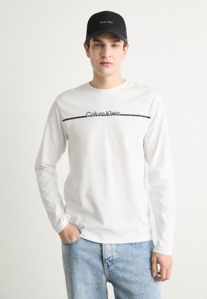 Hosszú ujjú fehér póló fekete "Calvin Klein" felirattal és vízszintes díszítéssel, világoskék farmernadrággal és fekete sapkával párosítva.