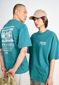 Teal cotton t-shirts with short sleeves; one features a white graphic on the back. Khaki cap and light grey shorts worn by the models.