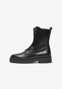 Botas de tornozelo em couro preto com acabamento liso, sola plana, design de atacadores e ilhós de metal. Possui uma alça na parte de trás.