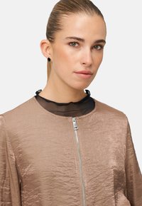 Lysebrun jakke med tekstureret finish, sølvlynlås og en sort netkrave ovenunder. Designet har en rund hals.