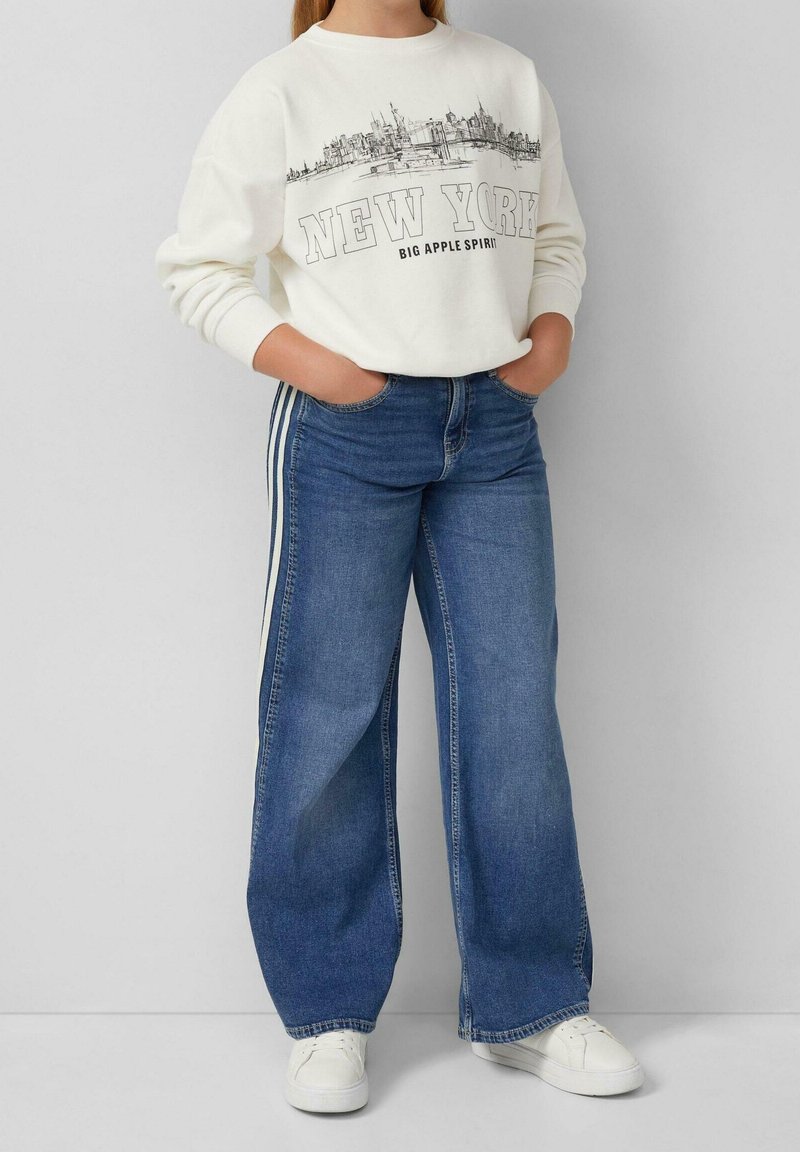 Personne portant un sweat-shirt blanc avec impression de la silhouette de New York, jean bleu à jambes larges avec bandes latérales, baskets blanches, debout les mains dans les poches.
