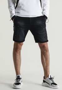 Pantalones cortos de mezclilla negra con una textura desvaída, bordes doblados y detalles desgastados. Llevados con zapatillas deportivas negras y grises.