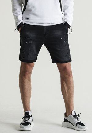 Pantalones cortos de mezclilla negra con una textura desvaída, bordes doblados y detalles desgastados. Llevados con zapatillas deportivas negras y grises.