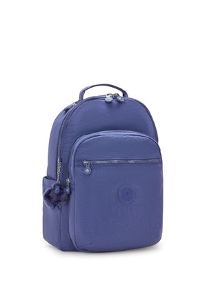 Purple nylon backpack with a rounded shape, front zip pocket, logo badge, and a fluffy charm attached to the zipper. Soft texture.
