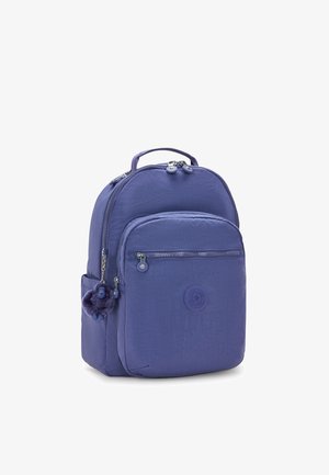 Purple nylon backpack with a rounded shape, front zip pocket, logo badge, and a fluffy charm attached to the zipper. Soft texture.