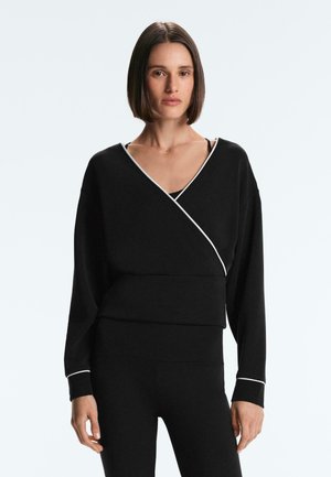 Vrouw met een zwarte wikkelstijl lange mouwen top en zwarte legging, staand tegen een effen lichte achtergrond.