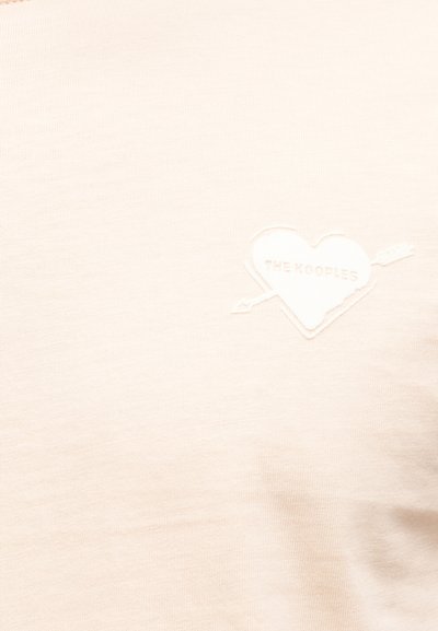 Ύφασμα εκρού με μπαλώμα σε σχήμα καρδιάς τρυπημένη από βέλος, κεντημένη με το κείμενο "THE KOOPLES" στη δεξιά πλευρά.