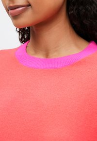Primer plano de una persona que lleva puesto un suéter de color coral con un cuello redondo acanalado de color rosa brillante y cabello oscuro rizado.