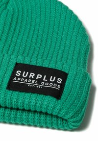 Zelena pletena kapa s rebrastom teksturom. Ima pravokutnu crnu naljepnicu s bijelim tekstom na kojem piše "SURPLUS APPAREL GOODS" i "EST. 1997."