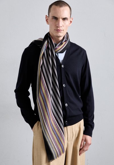 Schwarze Strickjacke mit Knopfleiste, kombiniert mit einem gestreiften Schal in kräftigen Farben und Mustern, lässig über einem hellen Hemd und beigen Hosen getragen.