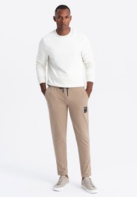 Ljust beige byxor med dragsko i midjan och en ficka i lappen. Kombinerat med en vit sweatshirt och grå sneakers. Slät textur genom hela plagget.