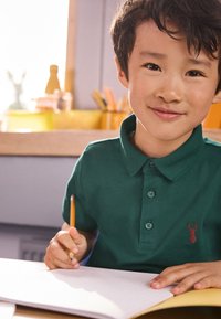 Grøn polo med knappet krave med et rødt hummel-emblem. Barn holder en blyant over en blank notesbog på et træbord.